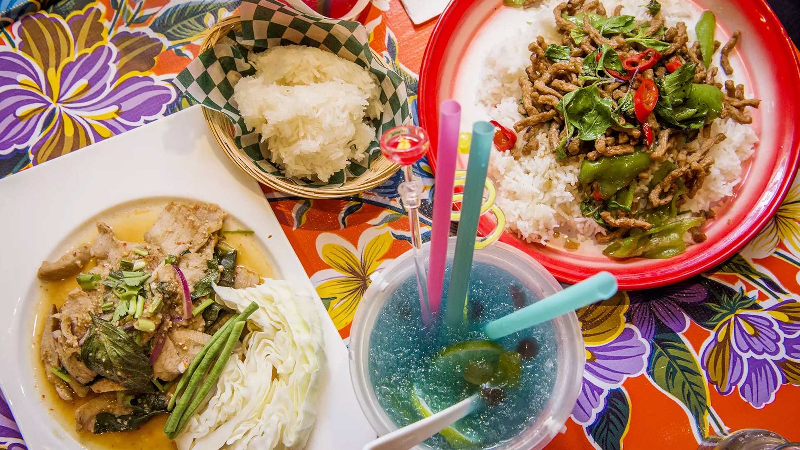 A table at SiLom: grilled pork jowl, holy basil beef over rice, sticky rice in a basket and a blue pea lemonade on a floral oilcloth